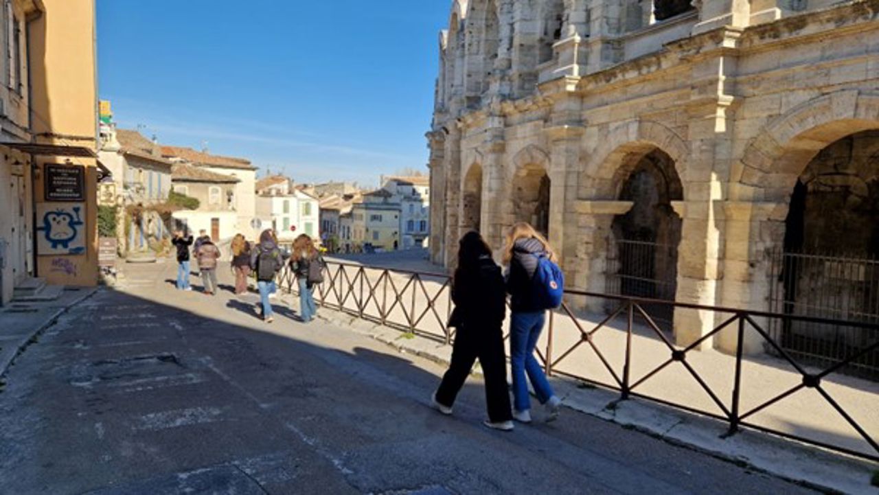 Ei gågate i den sørlege byen Arles i Frankrike. Det er solskin, og midt i bilete går det fleire menneske med ryggen til. Til høgre er det gamle, lyse steinbygninger med utsmykningar.