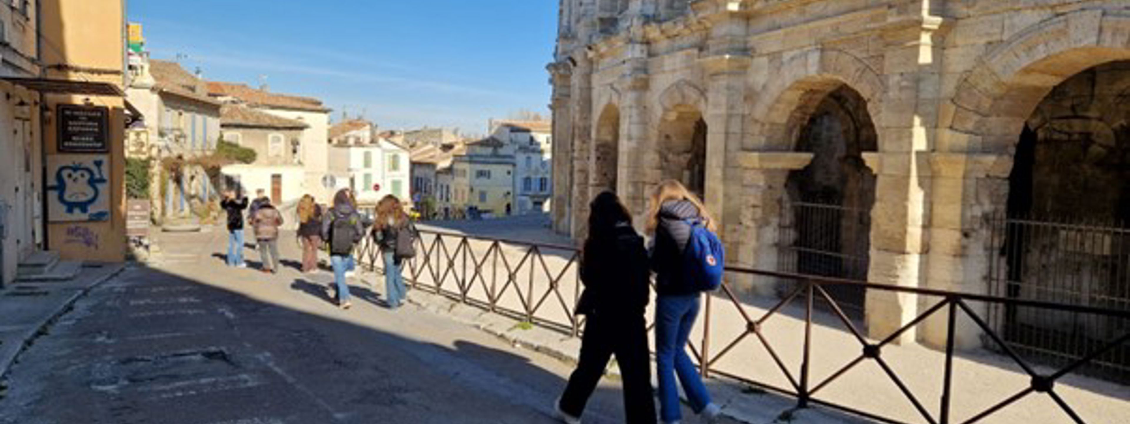 Ei gågate i den sørlege byen Arles i Frankrike. Det er solskin, og midt i bilete går det fleire menneske med ryggen til. Til høgre er det gamle, lyse steinbygninger med utsmykningar.