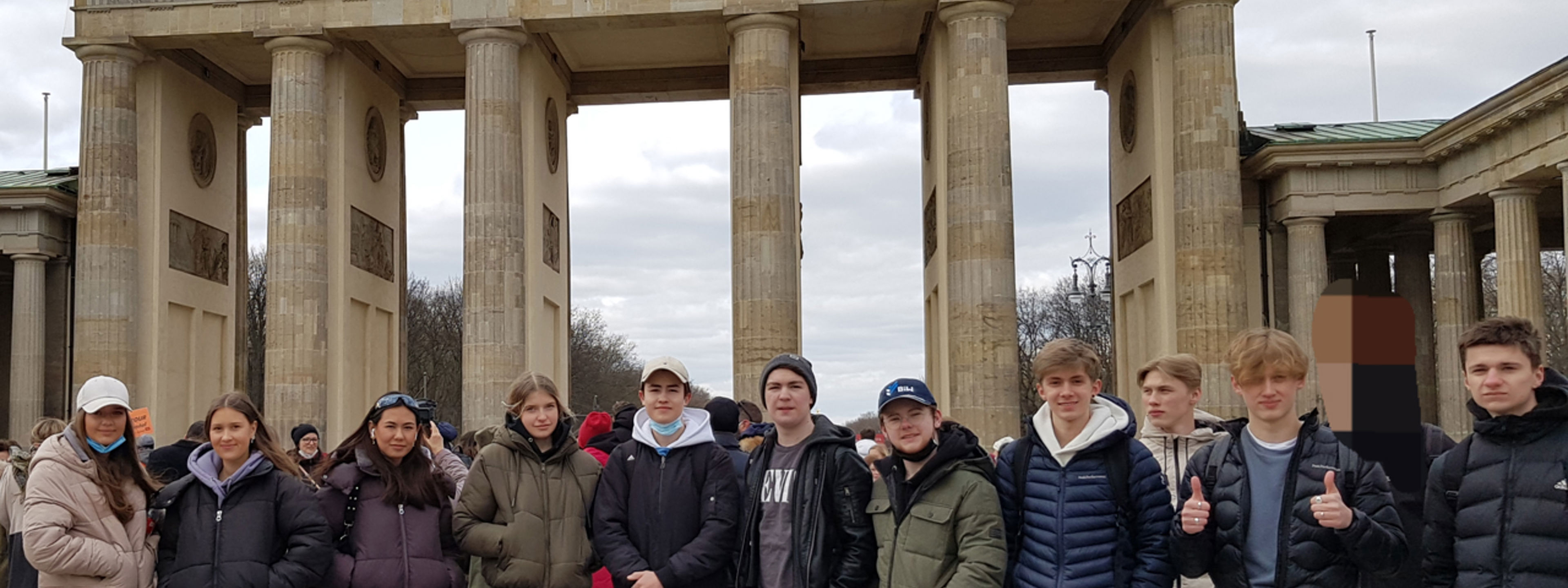 Elevar står på rekke framfor Brandenburger Tor. I bakgrunnen er ein stor byport i grå stein, med høge søyler. Øvst på monumentet er ei statue av ei vogn drive av fire hestar. I vogna sit ein figur med uniform. Figuren held eit septer med den tyske ørn og det prøyssiske Jernkorset